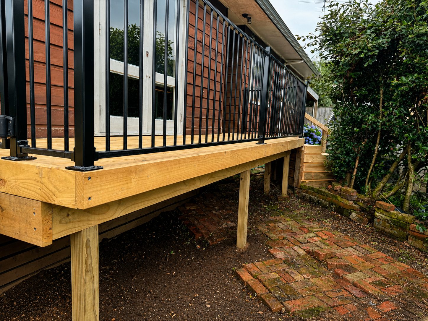 Elevated timber deck with black metal balustrade, built on posts over a brick path